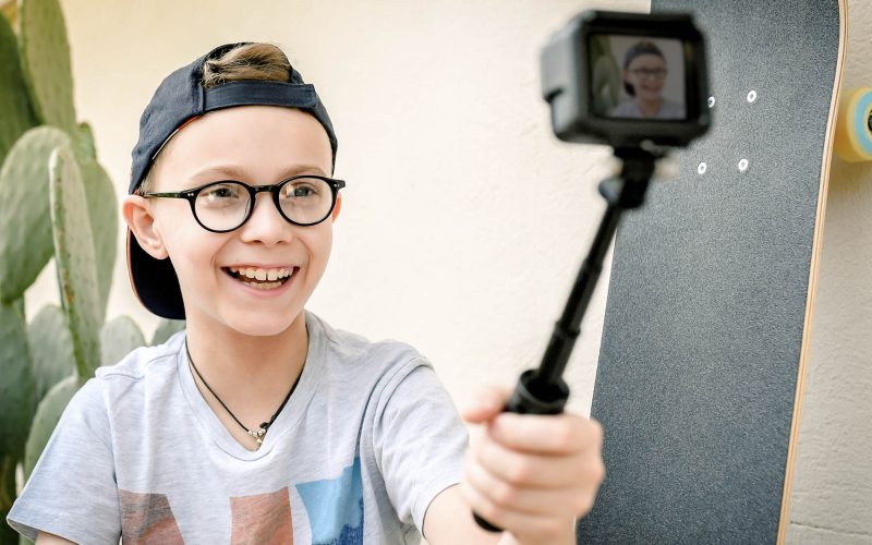 Young boy taking selfie on action camera outdoor with a skateboard and a cactus in background. Caucasian pre teen playing with action cam in the garden. Beautiful kid with hat and glasses smiling.; Shutterstock ID 1095383099; Purchase Order: -